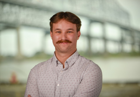 Smiling man with a mustache and crossed arms, blurred bridge in background.
