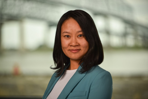 Professional Asian woman in a teal blazer, smiling, with a blurred outdoor background.