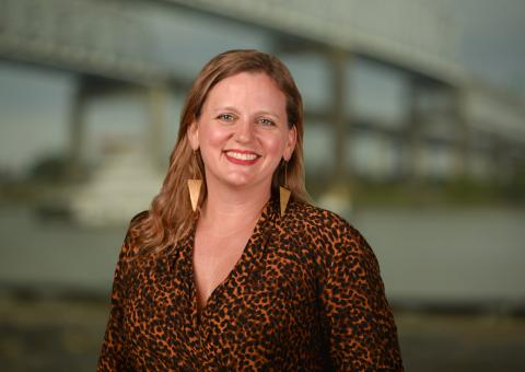 Smiling woman in patterned top with long brown hair, blurred bridge in background.