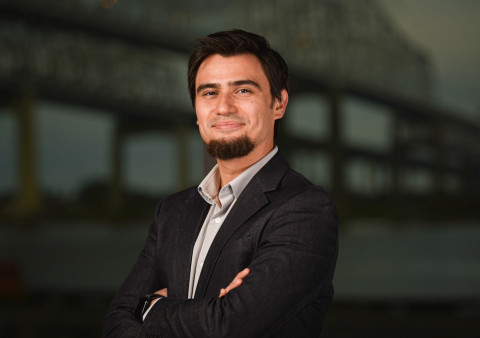 Smiling man with a dark beard and suit, arms crossed, blurred industrial background.