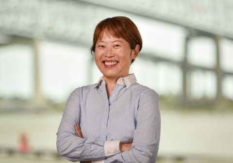 Smiling woman with short brown hair, blue striped shirt, arms crossed. Blurred background.