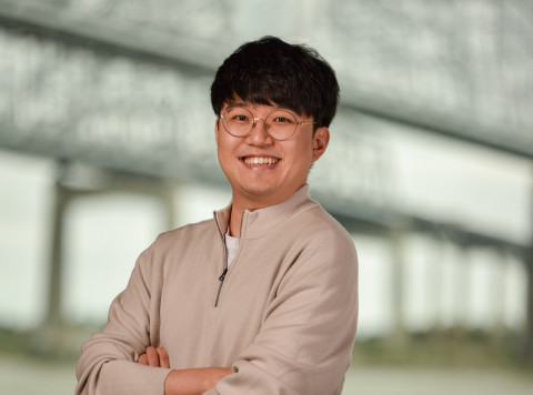 Cheerful man in beige quarter-zip and glasses, arms crossed, blurred bridge background.