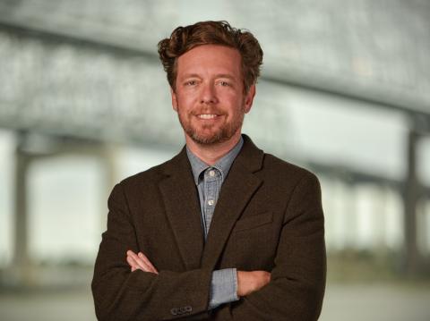 Smiling man with reddish hair and beard, wearing a brown jacket, arms crossed.