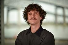 Young man with curly hair and mustache smiling subtly.