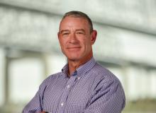 Headshot of a smiling man in a purple checkered shirt, arms crossed.
