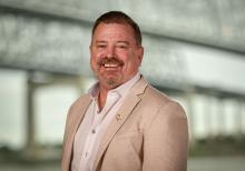 Smiling man in a beige jacket against a blurred bridge background.