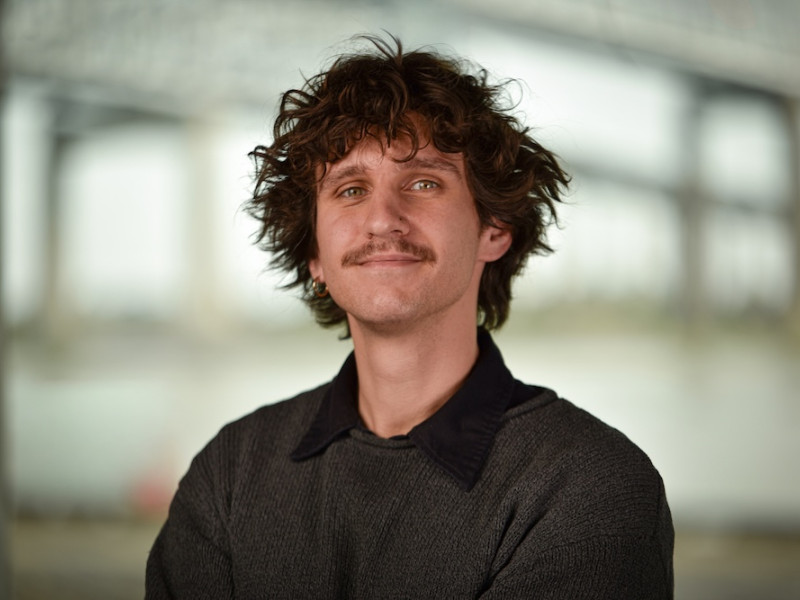 Young man with curly brown hair, mustache, and dark sweater, smiling faintly.