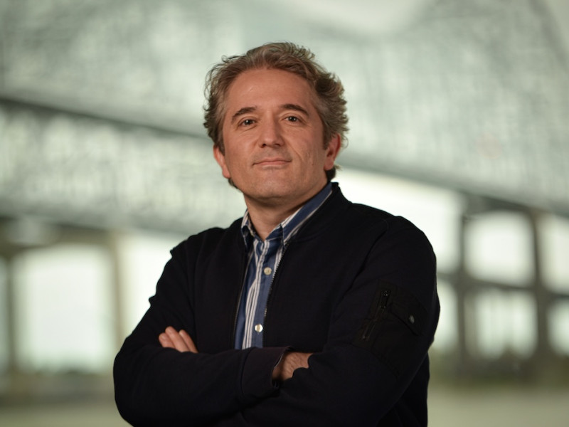 Man with graying hair, blue striped shirt and navy cardigan, arms crossed.