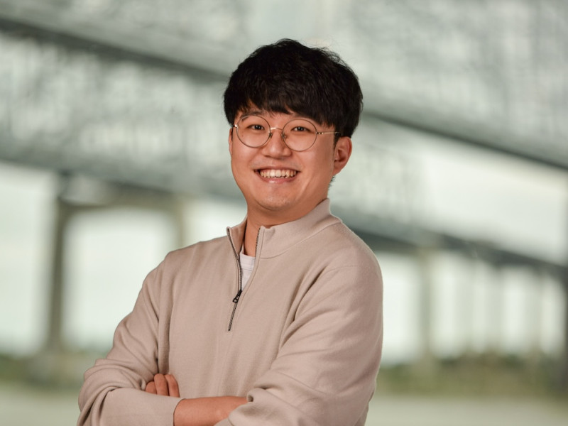Cheerful man in beige quarter-zip and glasses, arms crossed, blurred bridge background.