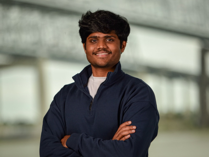 Smiling young man with dark skin and hair, wearing a dark blue quarter-zip, arms crossed.