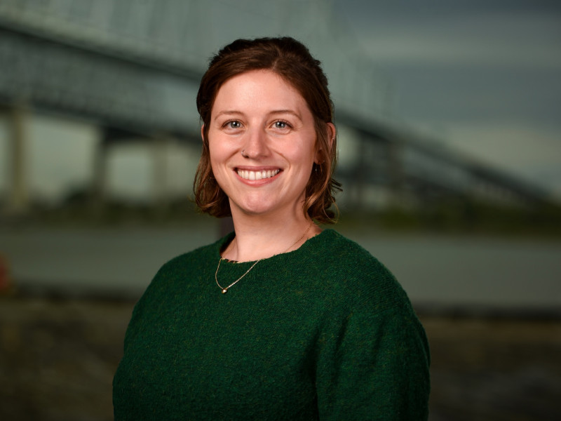 Smiling woman in a dark green sweater outdoors, with a blurred bridge behind her.